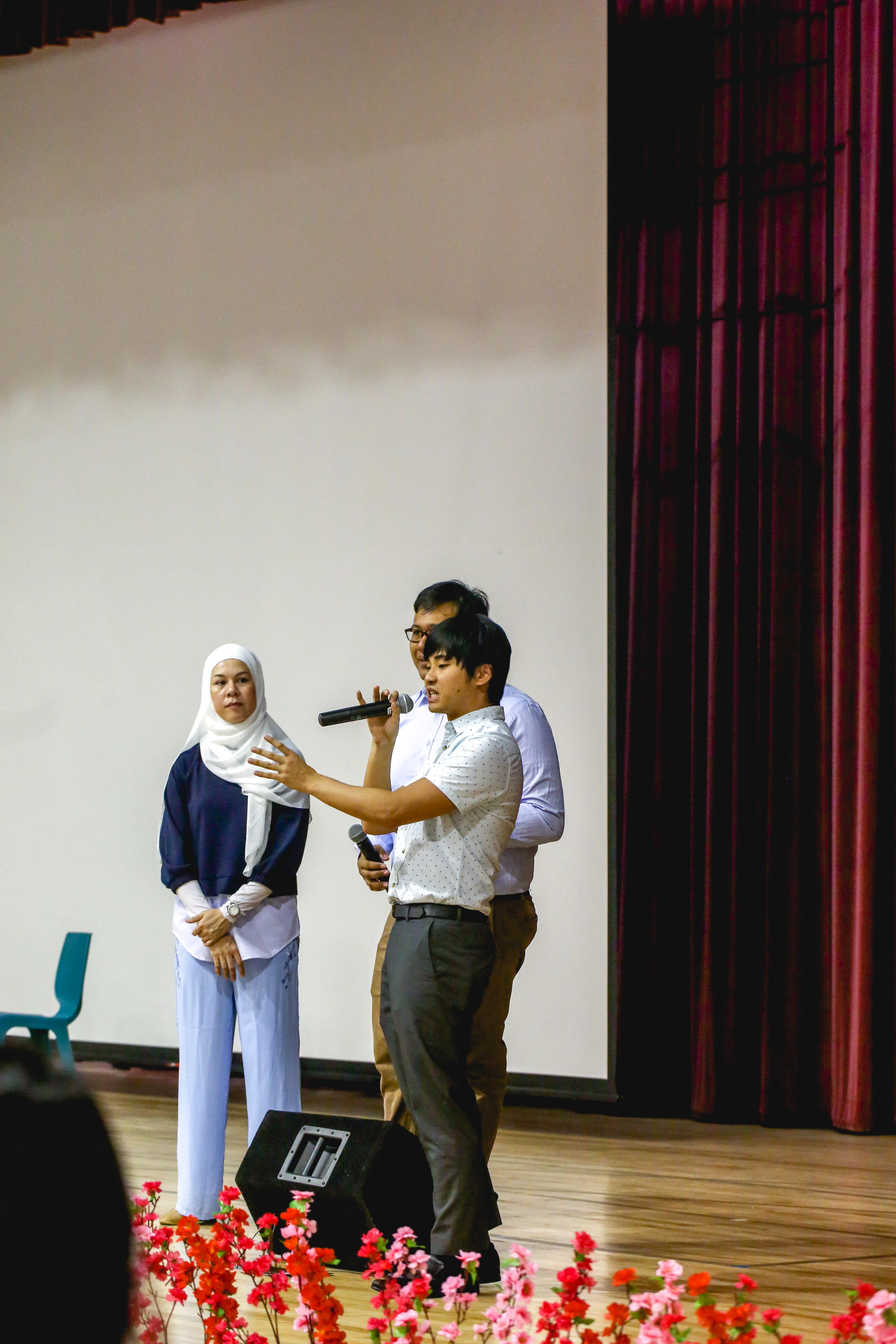 A young man speaks into a microphone on stage with two others nearby, flowers in the foreground and a red curtain behind.
